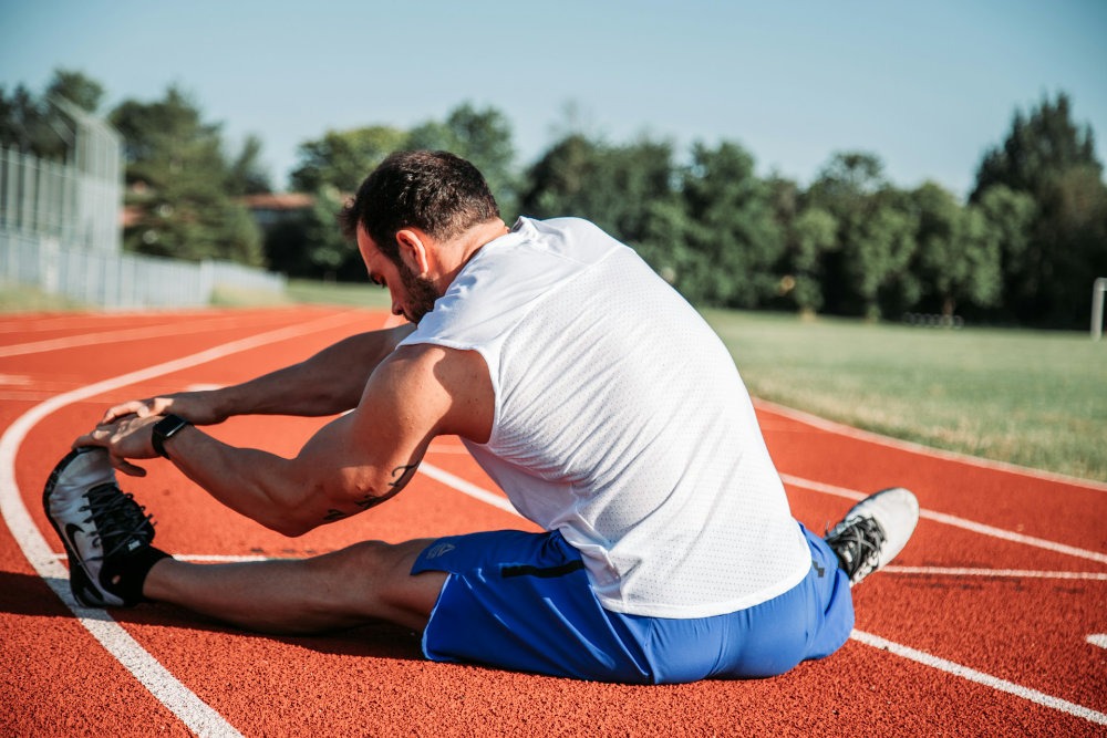 Homme qui s'étire les jambes sur une piste d'athlétisme. Il est assis de dos.
